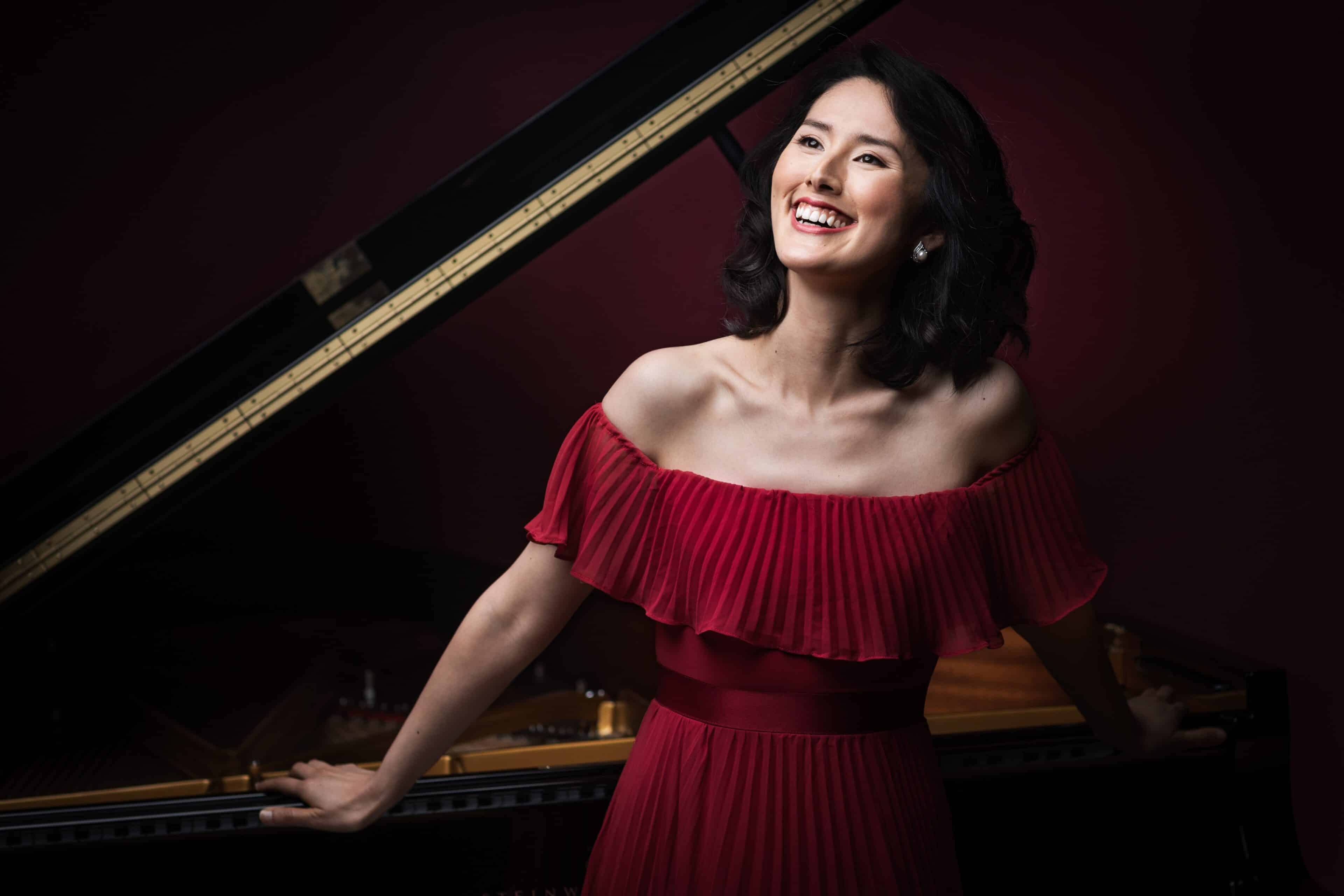 Photo of Yuko Sano, wearing a red dress and smiling in front of an open grand piano